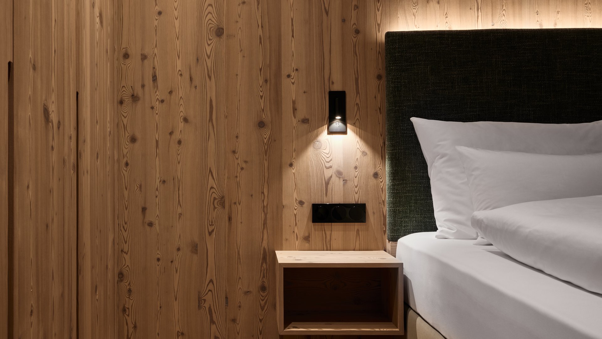 Frühbucher Bonus & Bestprice in Meransen / Südtirol | Mountain Lodge Margit Detailansicht eines Schlafzimmers in der Mountain Lodge Margit mit einem gemachten Bett, weißer Bettwäsche, dunkelgrünem Kopfteil und einer Wandverkleidung aus natürlichem Lärchenholz mit Nachttisch.